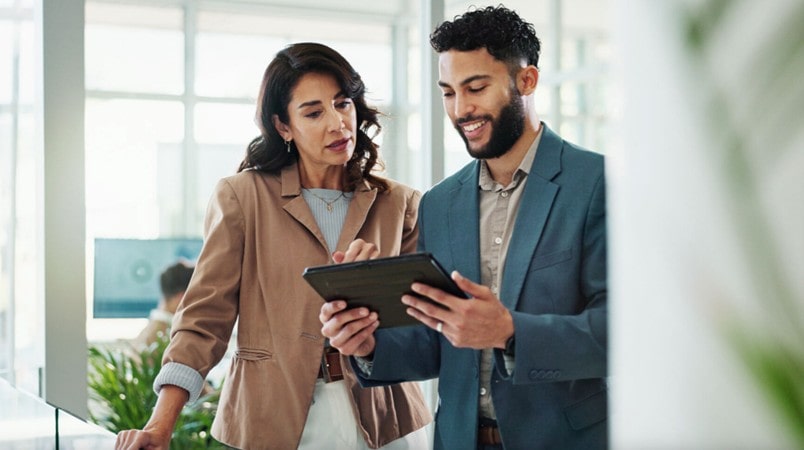 Two people standing together reviewing information on a digital tablet in an office setting.