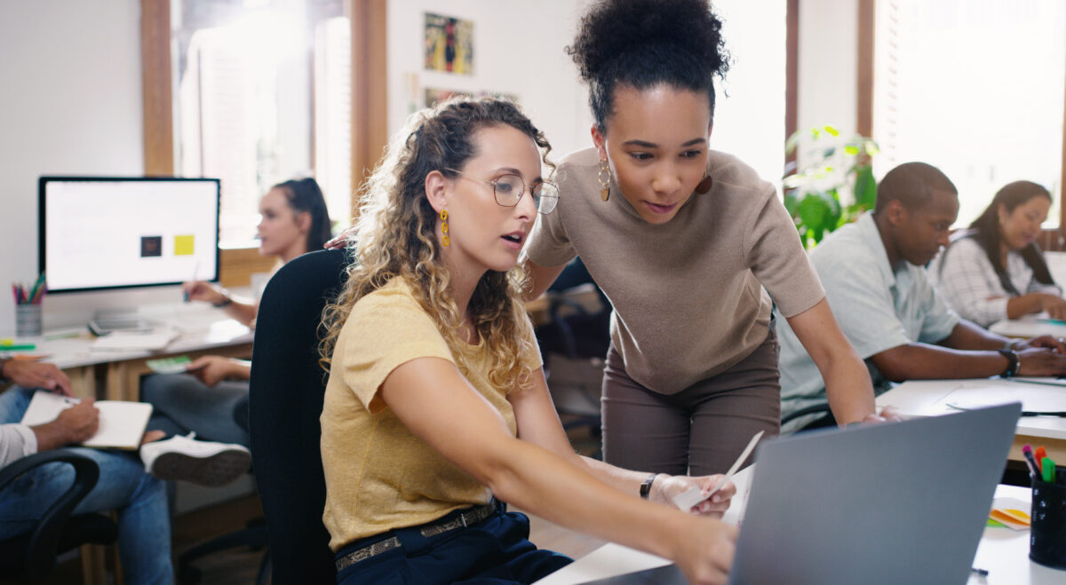 Two business women talking at a work table and looking at a laptop.